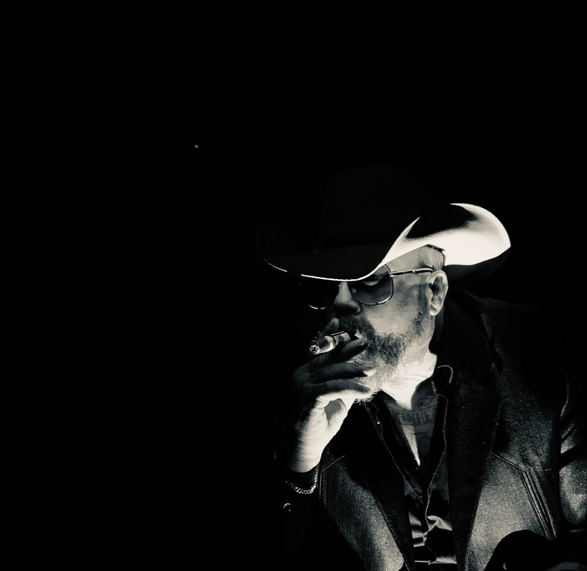 Band portrait in black and white — a figure in a cowboy hat and dark glasses smoking a cigar, lit hard against black